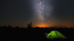 Een nachthemel met sterren en de Melkweg, een bergrug in silhouet, en een groene tent.