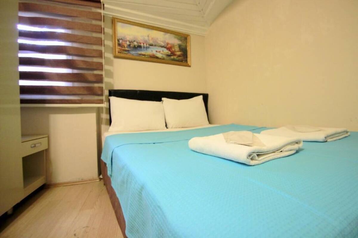 A neatly made bed with a blue bedspread and white pillows, a painting on the wall, and a window with blinds.
