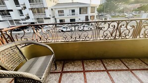 A balcony with a wicker chair, a metal railing, and a view of a cityscape with buildings and parked cars.