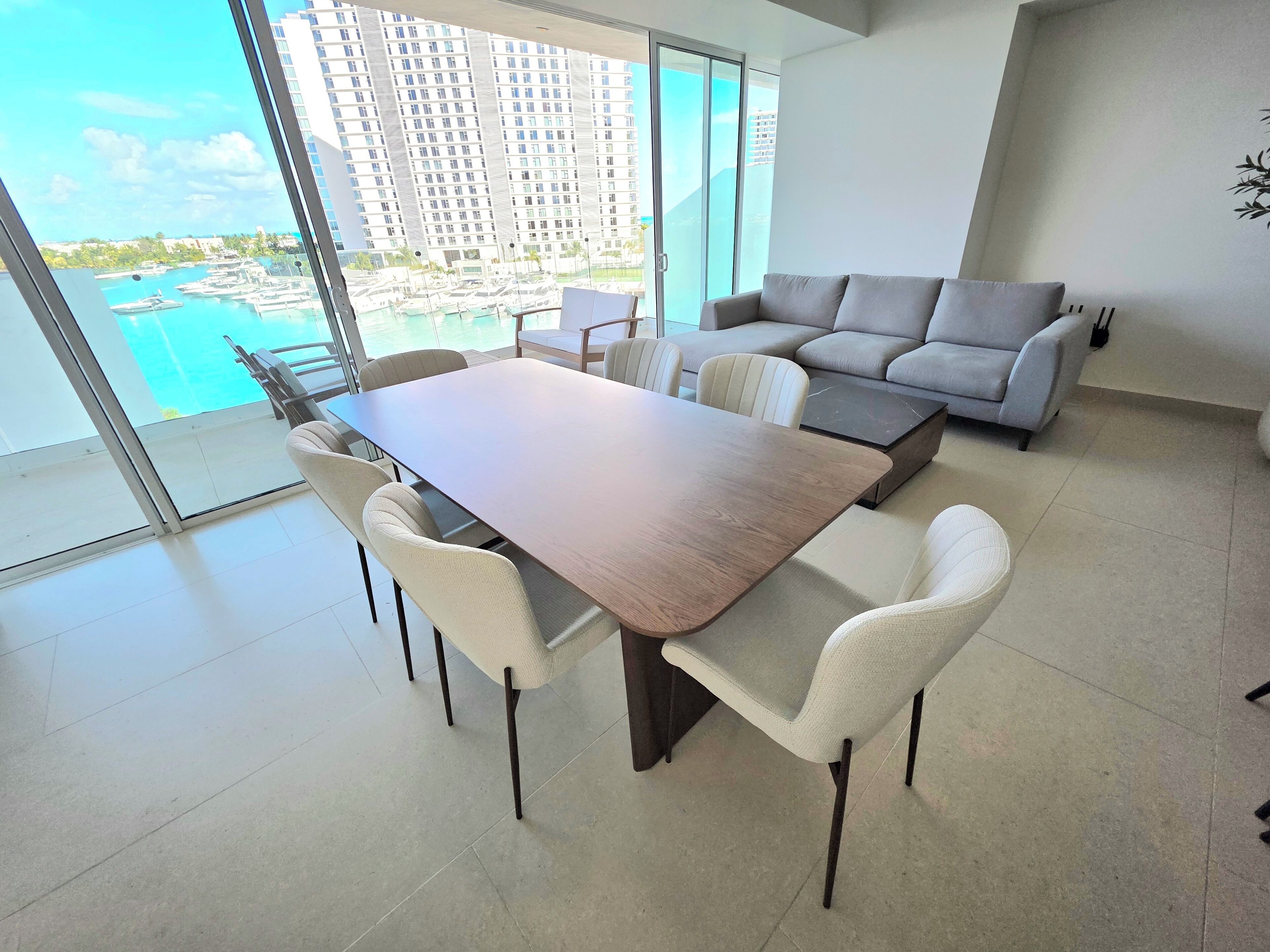 A modern living room with a dining table and sofa, large windows with a view of a marina and buildings.
