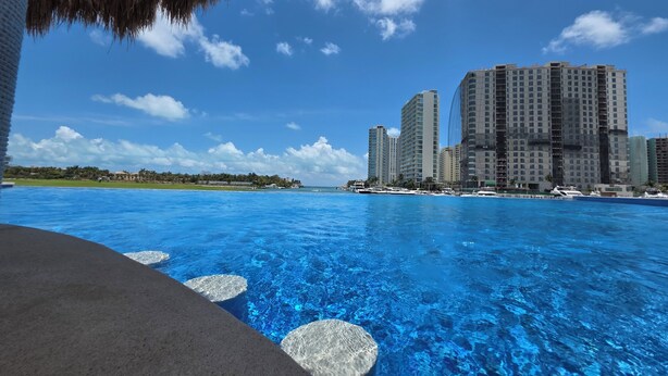 A clear view of a swimming pool with a thatched roof structure above, surrounded by tall residential buildings and a clear blue sky. - Marea condo vista espectacular 4pax (Cancun)
