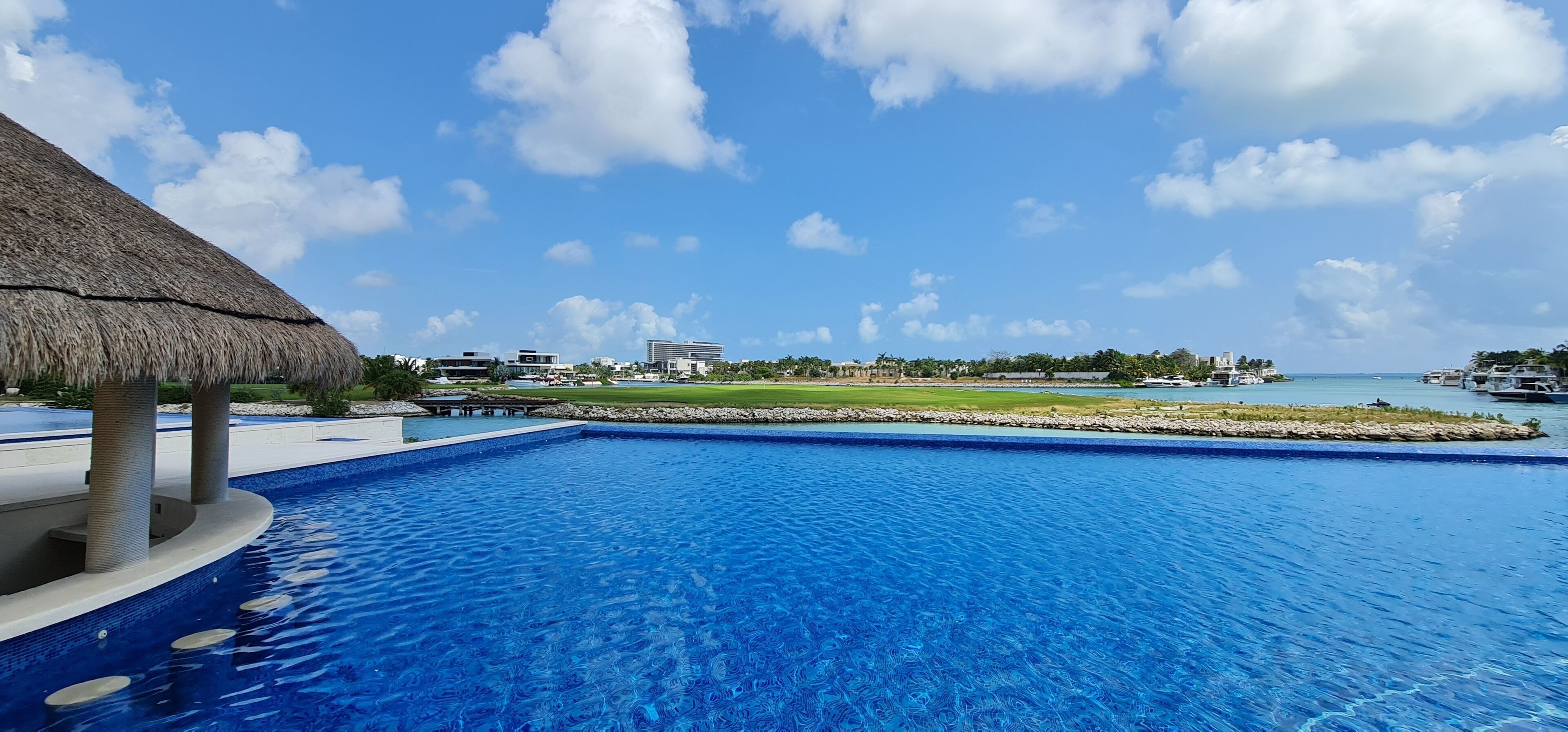 Une piscine dotée d’une structure avec toit de chaume, une vue sur un port de plaisance avec des bateaux, et un ciel bleu dégagé.