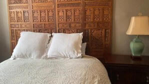Una cama con una colcha estampada y dos almohadas, un cabecero de madera con intrincados grabados, y una lámpara verde sobre una mesita de noche.
