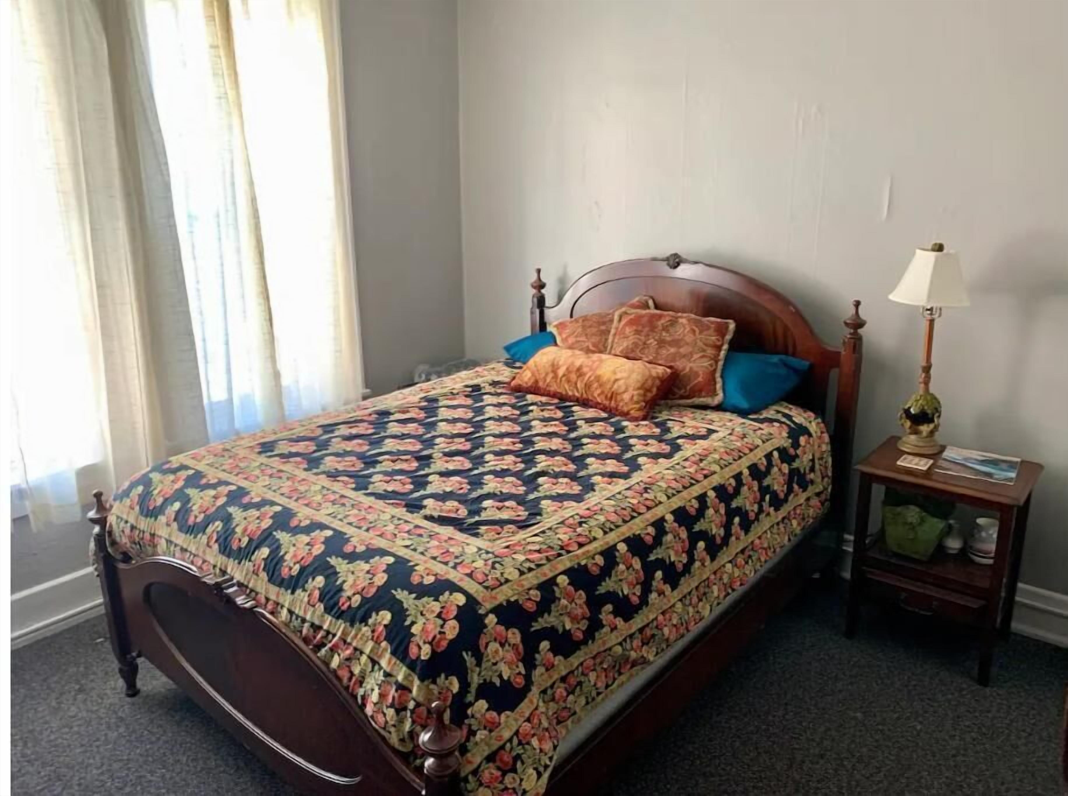A neatly made bed with a floral-patterned bedspread, a wooden headboard, and a bedside table with a lamp and a small decorative item.