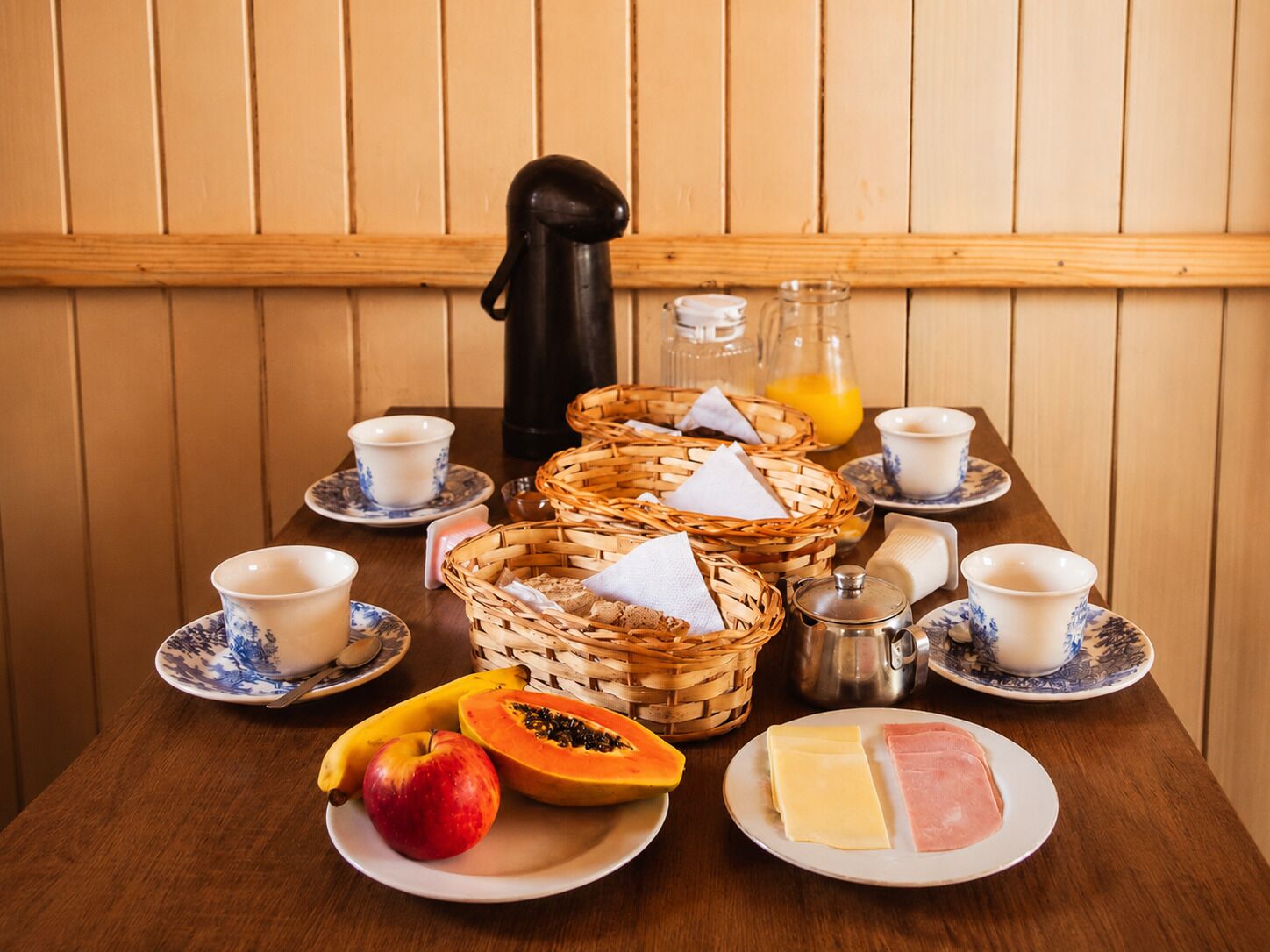 A breakfast table with plates of food, cups, a kettle, and a thermos.