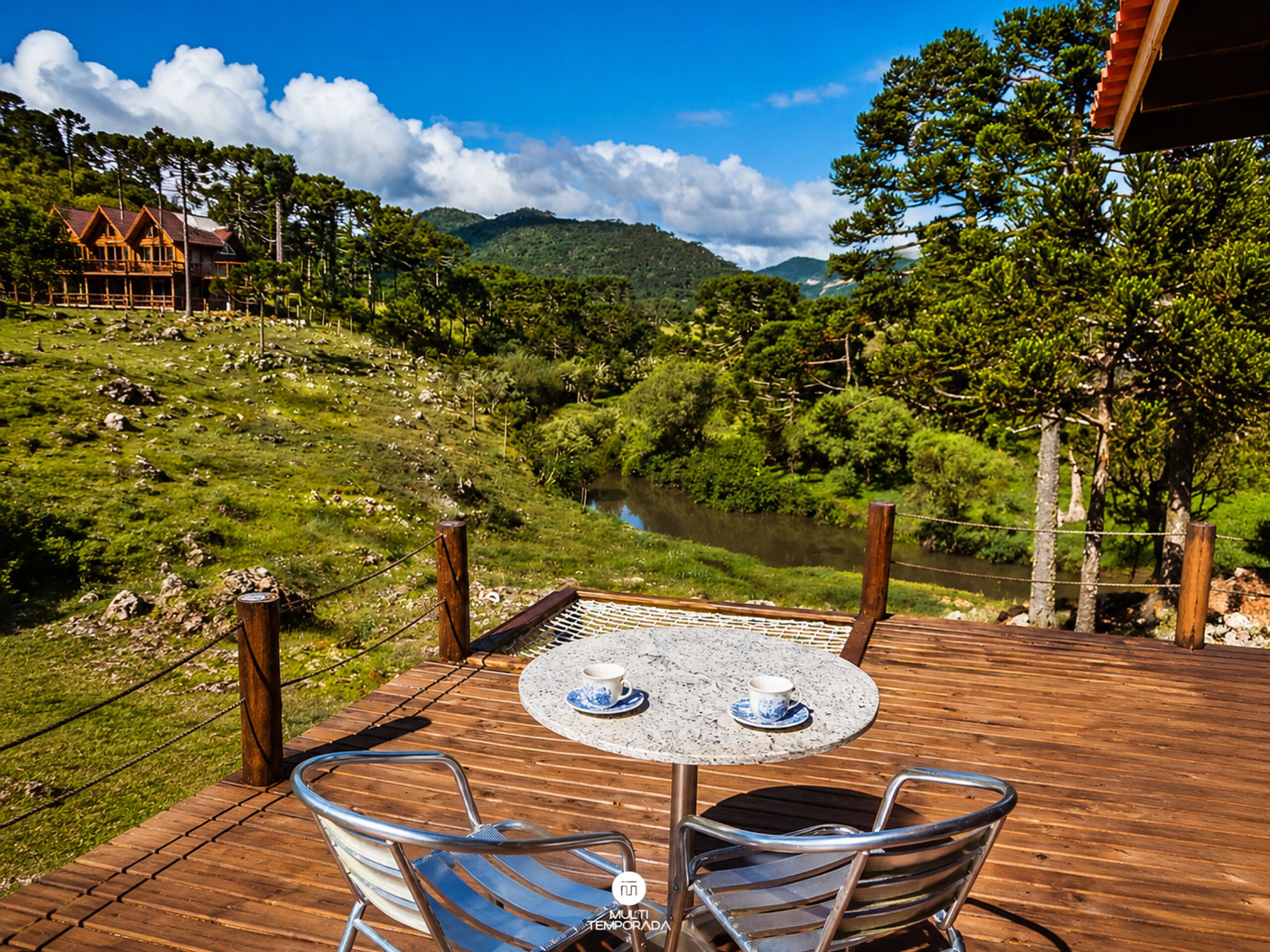 Terraza de madera con una mesa y sillas con vista a un río y colinas boscosas.