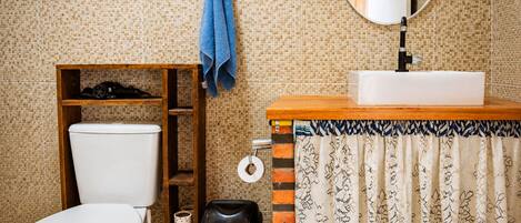 Une salle de bain comprenant des toilettes, une étagère avec des articles de toilette, une poubelle, une serviette accrochée au mur et un miroir.