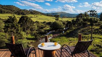 A wooden deck with a round table and two chairs, overlooking a lush valley with a river and distant hills.
