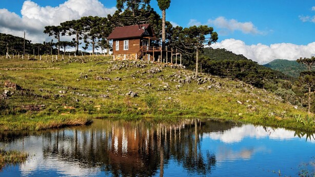 A wooden cabin with a red roof, surrounded by a grassy landscape and a body of water reflecting the sky. - Recanto das Águas - Urubici - SC (Urubici)
