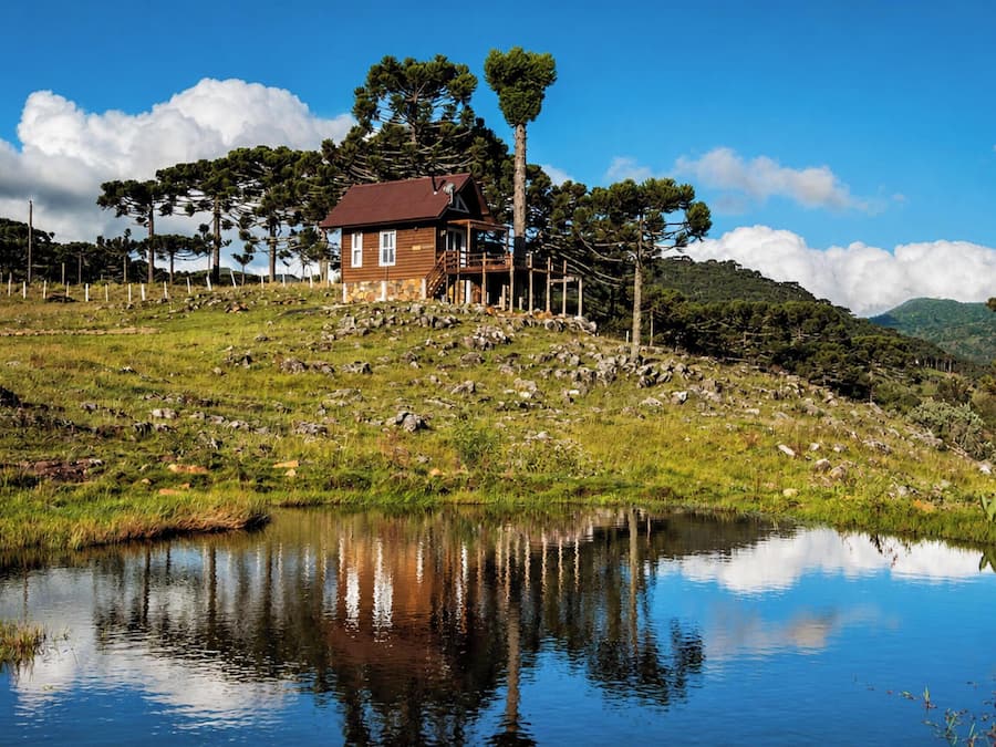 A wooden cabin with a red roof, surrounded by a grassy landscape and a body of water reflecting the sky.