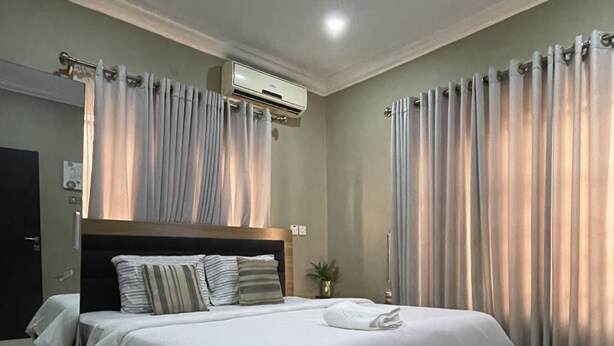 A modern bedroom with a large bed, a bedside table, a wall-mounted air conditioner, and a window with curtains. - NUMBER ELEVEN APARTMENTS (Lagos)