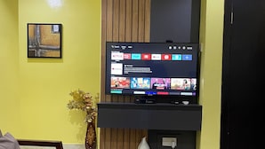 A living room with a flat-screen TV mounted on a wooden panel wall, a grey sofa, a coffee table, and a vase with flowers. - NUMBER ELEVEN APARTMENTS (Lagos)