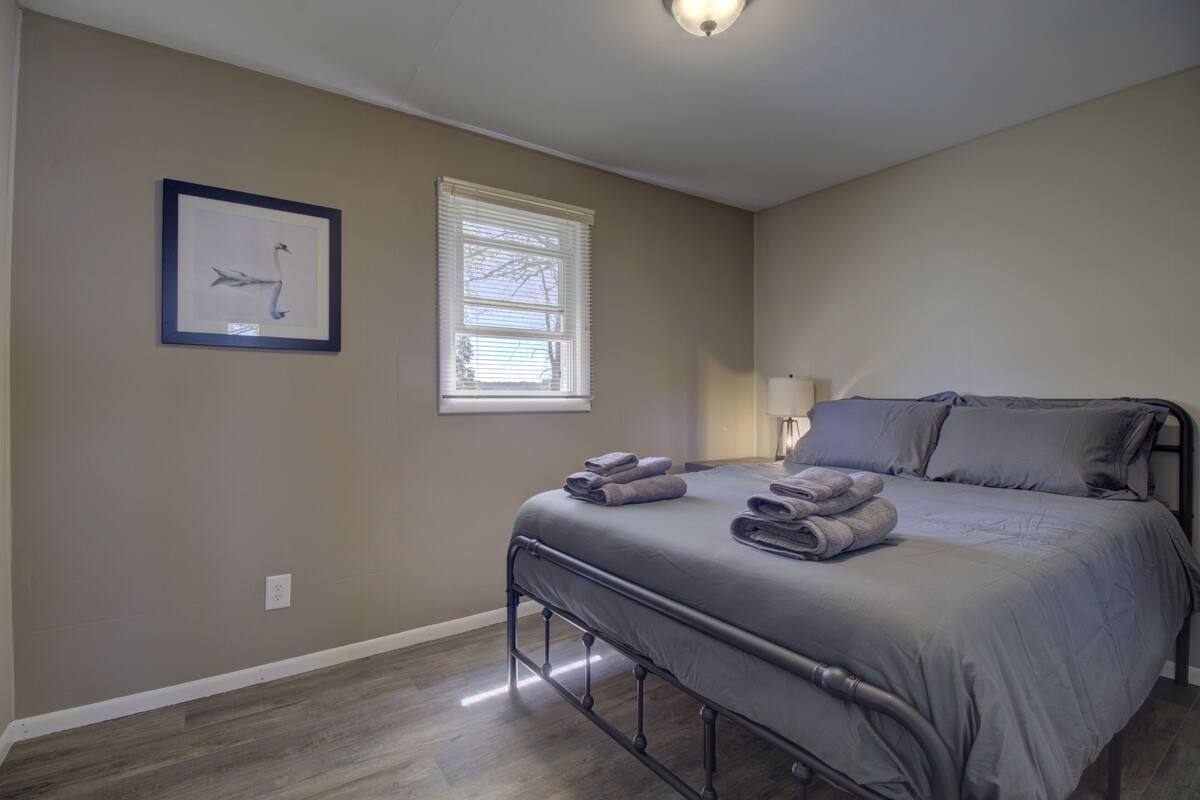 A bedroom with a bed, a window with blinds, a wall-mounted light fixture, and a framed picture on the wall.