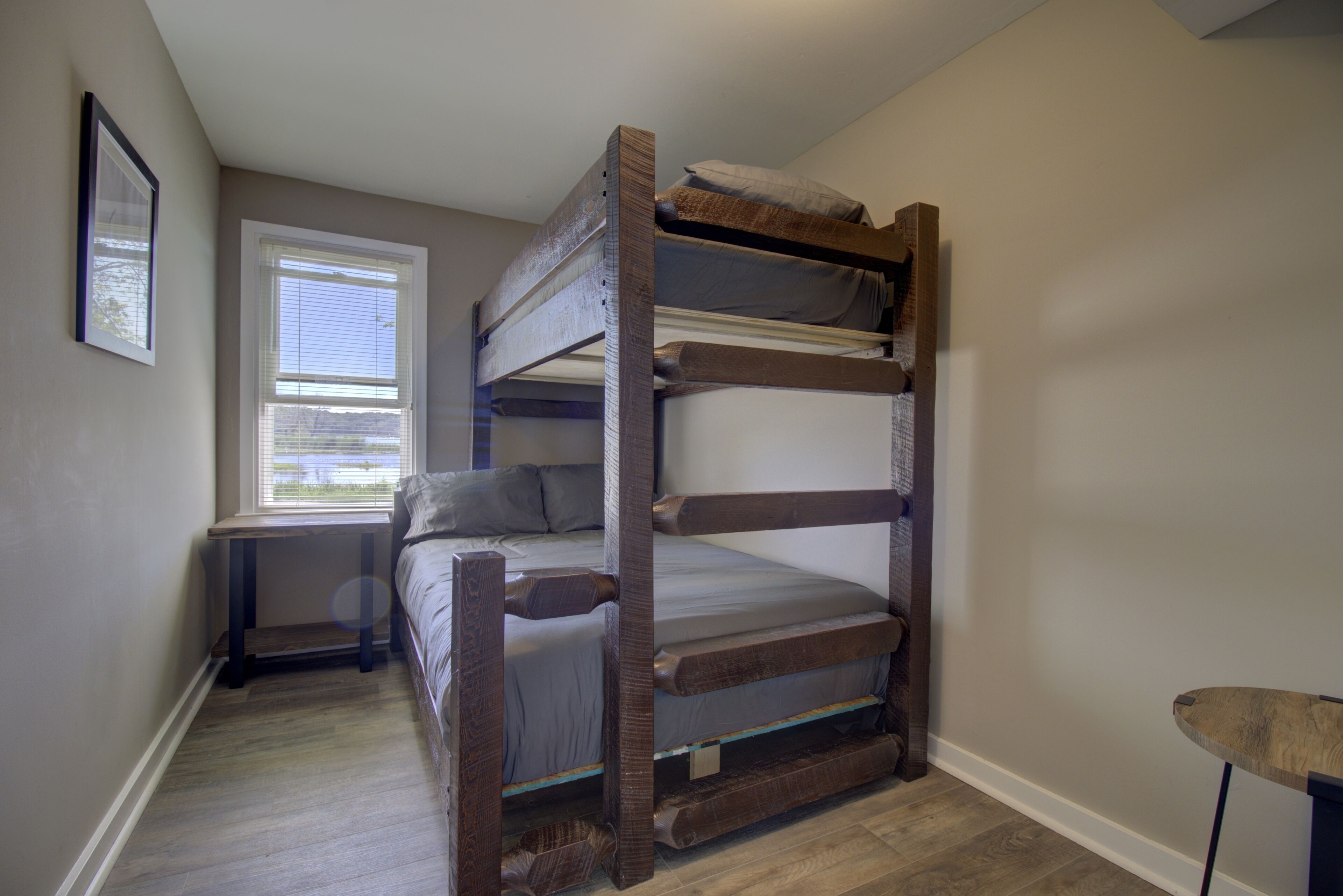 A bunk bed in a room with a window, a small table, and a framed picture on the wall.