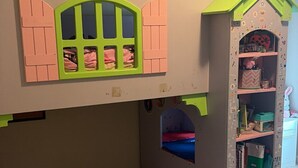 Una casita de juegos para niños con techo verde, contraventanas rosas y un ventanal pequeño.