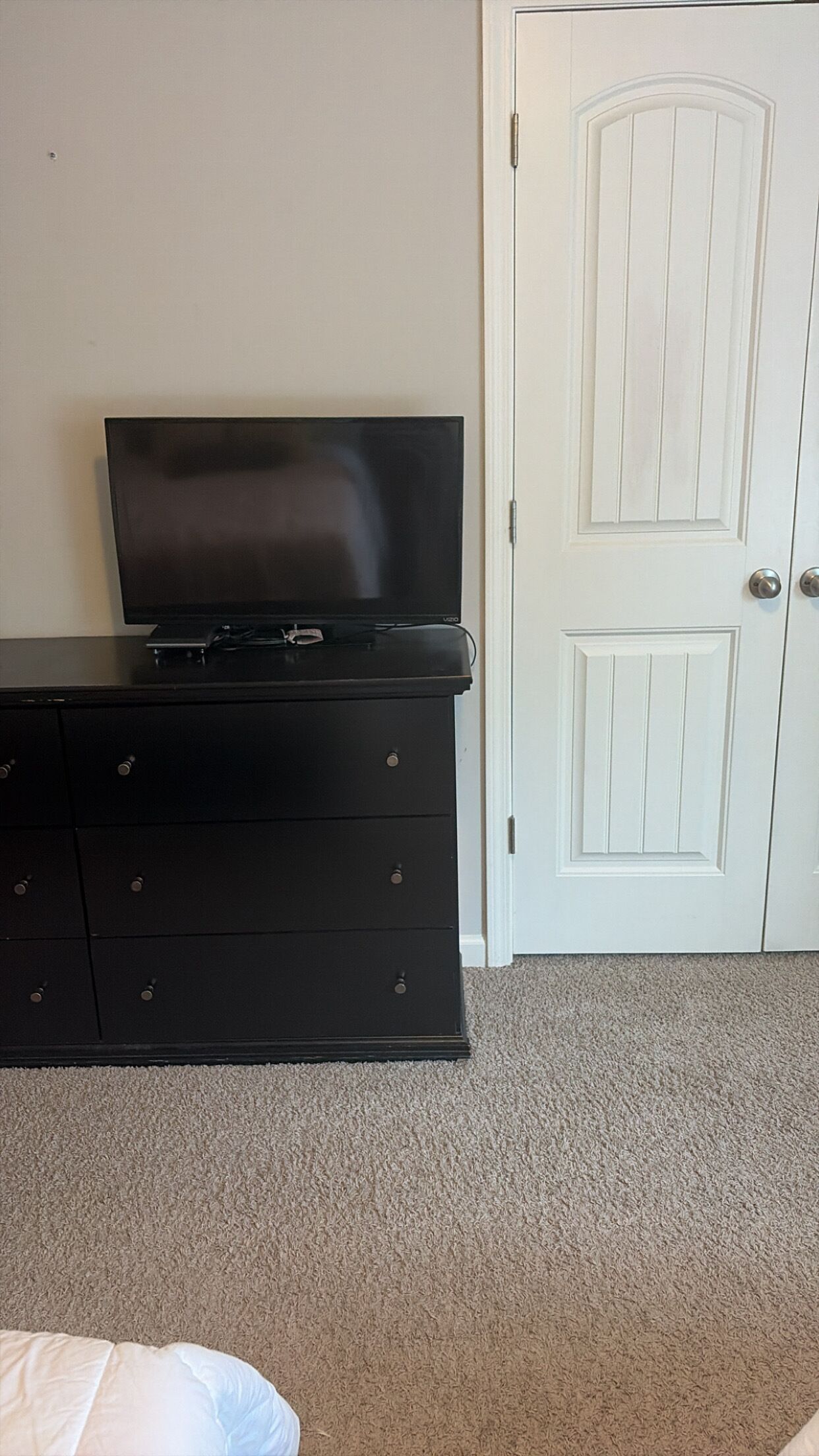 A room with a black dresser, a television on top, a white door, and a carpeted floor.