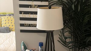 Una mesita de noche con una lámpara, un adorno con rayas en la pared y una planta grande en maceta.