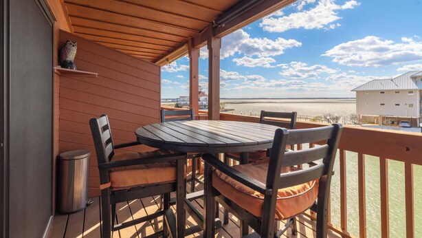 Een houten terras met een tafel en stoelen, met uitzicht op een strand en een wit gebouw.