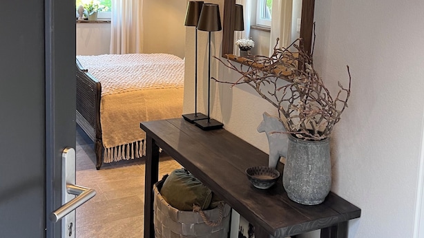 Habitación de hotel con cama, mesita de noche con lámpara, florero con ramas y espejo en la pared.