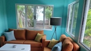 Een woonkamer met een bank, een bijzettafeltje en een lamp. Er zijn ramen met uitzicht op groen.