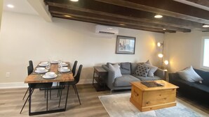 Un salon moderne comprenant un coin repas, un canapé gris, une table basse en bois et un plafond avec des poutres apparentes.