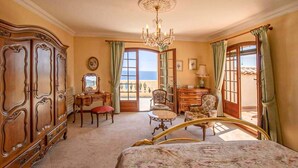 A room with a bed, a dresser, a desk, and a chair. There is a chandelier and a view of the sea through a window.