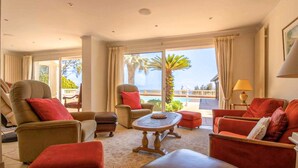 A spacious living room with a large window offering a view of the sea and palm trees. There are several comfortable seating options, including a sofa and armchairs.