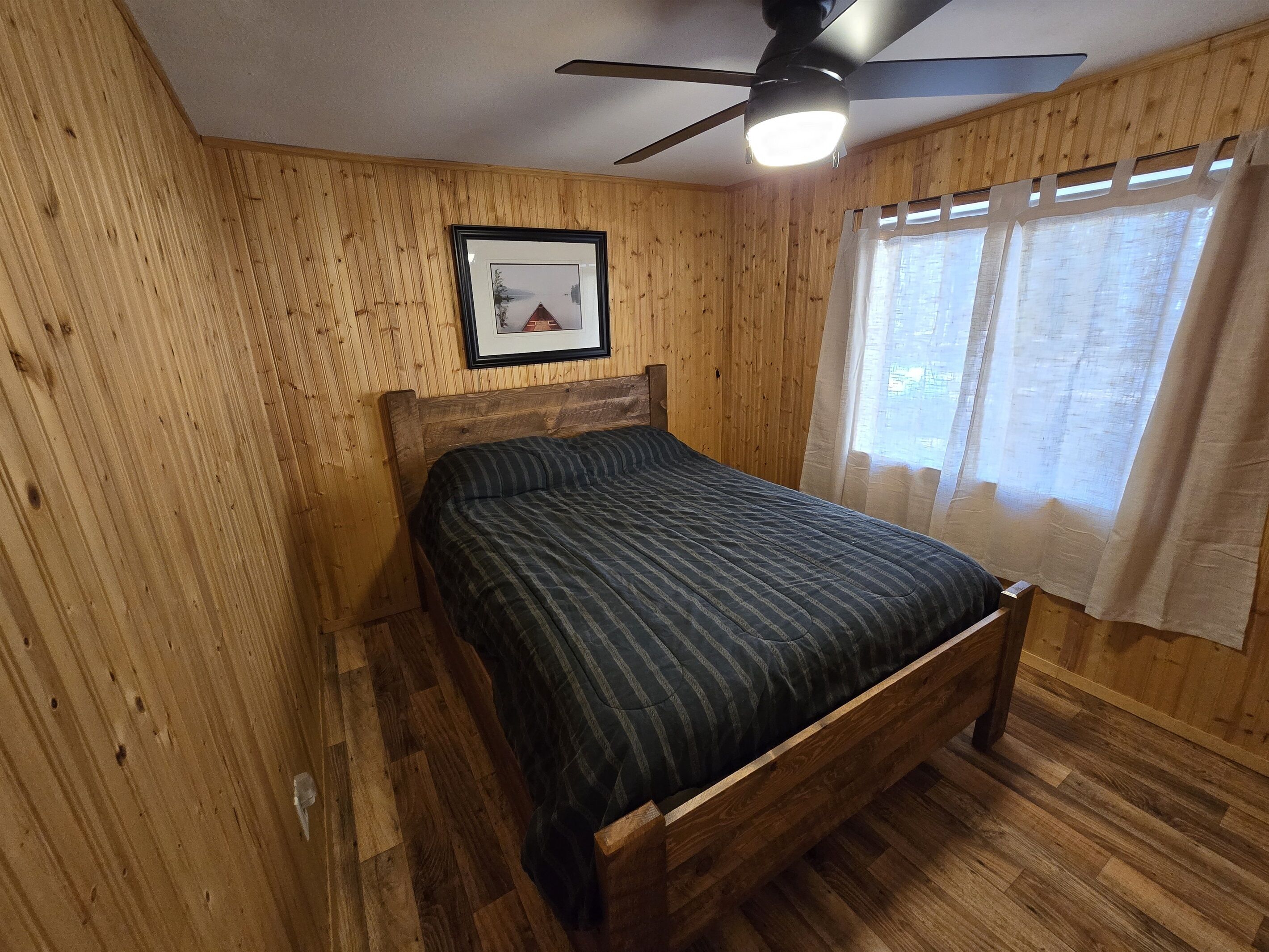 A wooden-paneled room with a bed, a ceiling fan, and a window with curtains.