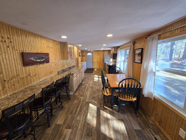 A kitchen with wooden walls, a granite countertop, and a dining area with a wooden table and chairs. - Somo River Home - Fire Pit, Dog Friendly, W&D, Kayaks, Canoe (Tomahawk)