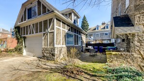 Une maison à deux étages avec un garage, des murs en pierre et une cour entourée d’une clôture, aménagée avec du mobilier d’extérieur.