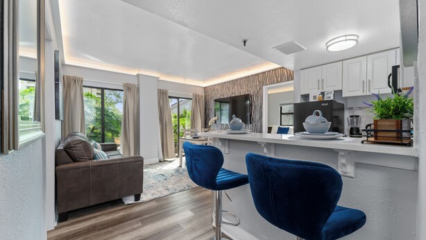 Eine moderne Küche mit weißen Schränken, einem schwarzen Kühlschrank und einer Kaffeemaschine. Es gibt einen Essbereich mit einem braunen Sofa und einem blauen Barhocker.