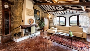 A rustic living room with exposed wooden beams, a stone wall, a fireplace, a sofa, and large windows offering a view.