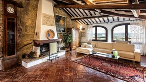 A rustic living room with exposed wooden beams, a stone wall, a fireplace, a sofa, and large windows offering a view.