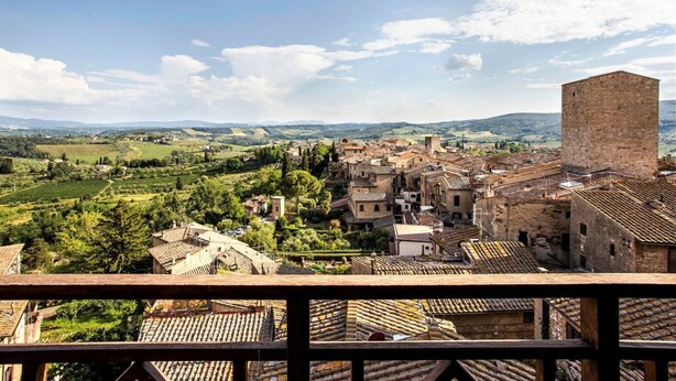 A historic town with terracotta-roofed buildings, a prominent tower, and rolling hills in the background.