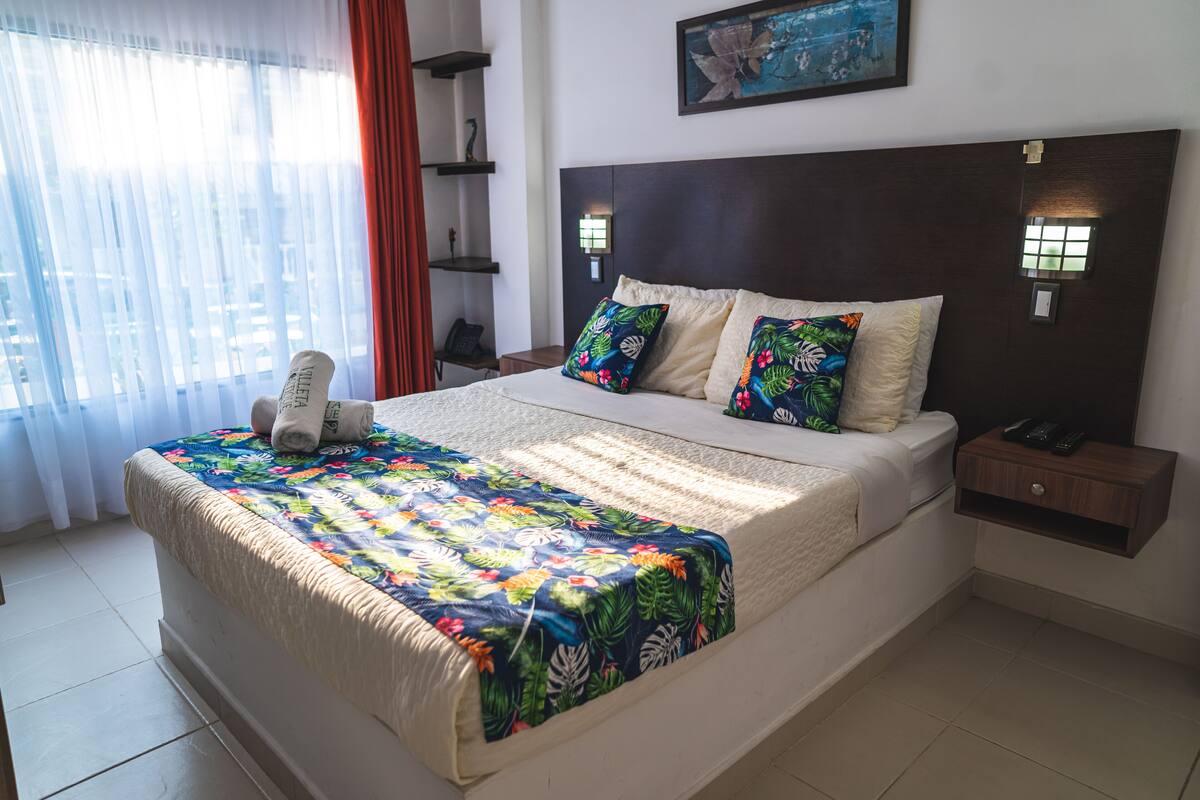 Una cama bien tendida con una colcha floral, un cabecero con luces empotradas en la pared y una mesita de noche con un teléfono y controles remotos.