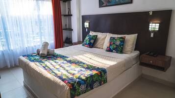 A neatly made bed with a floral bedspread, a headboard with wall-mounted lights, and a small bedside table with a phone and remote controls.