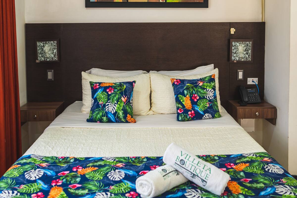 Habitación de hotel con cama, dos almohadas, una colcha, un cuadro, aire acondicionado, teléfono y mesita de noche.