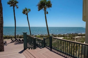 A wooden deck with a railing, overlooking a beach with palm trees and the ocean.