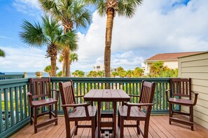 A wooden deck with a table and chairs, overlooking a coastal area with palm trees and a view of the ocean.
