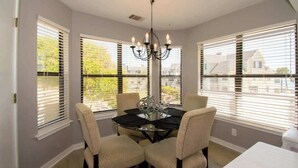 Une salle à manger avec une table en verre, quatre chaises, un lustre et de grandes fenêtres équipées de stores.
