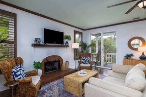 A living room with a fireplace, television, wicker chairs, a sofa, and a view of the outdoors.