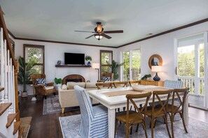 Amplia sala de estar con chimenea, ventilador de techo y comedor con sillas de madera y una mesa blanca.