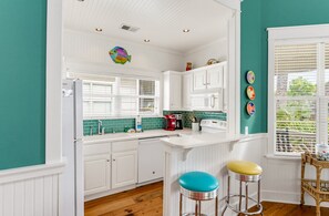 A kitchen with white cabinets, a white countertop, and a window with blinds. There are two stools with colorful seats.