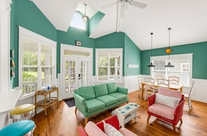 A room with a green sofa, a red chair, a dining table with chairs, a ceiling fan, and a window with blinds.