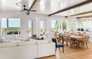 Ampla sala de estar com sofá branco, mesa de jantar e cadeiras, com grandes janelas que oferecem vista para o exterior.