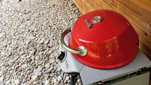 A red barbecue grill with a digital thermometer, placed on a gravel surface in front of a wooden house with large windows and a wooden ceiling.