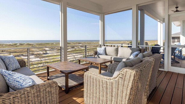 Un espace extérieur abrité, aménagé avec des meubles en osier, une table en bois et de grandes fenêtres offrant une vue sur la plage et les dunes.