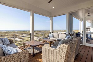 A covered outdoor area with wicker furniture, a wooden table, and large windows offering a view of the beach and dunes. - 711 Yellow Perch Road (Fripp Island)