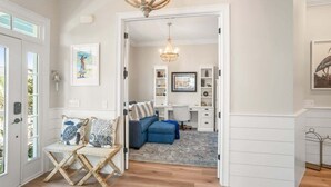 A hallway with a wooden floor, a blue sofa, a white desk, and a chandelier. - 778 Marlin Drive (Fripp Island)