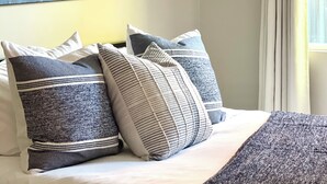 Ein Bett mit weißer Bettwäsche und mehreren Kissen, ein Gemälde mit Löwenzahn an der Wand und ein Fenster mit Vorhängen.